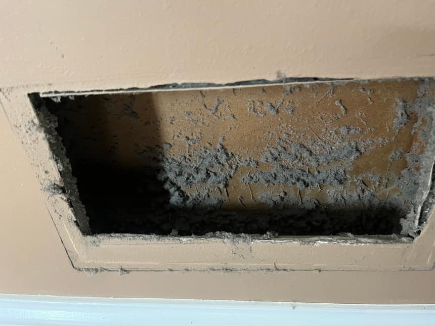 An open wall return with the grille off, the duct behind it furred over in grey dust