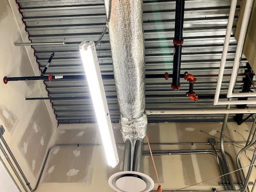 Insulated supply ductwork running under an open commercial ceiling deck alongside sprinkler and conduit runs