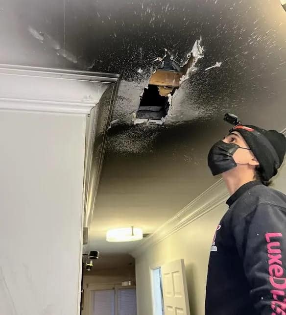 A Luxe Ducts technician in a respirator looking up at a smoke damaged hallway ceiling, burned open around the register