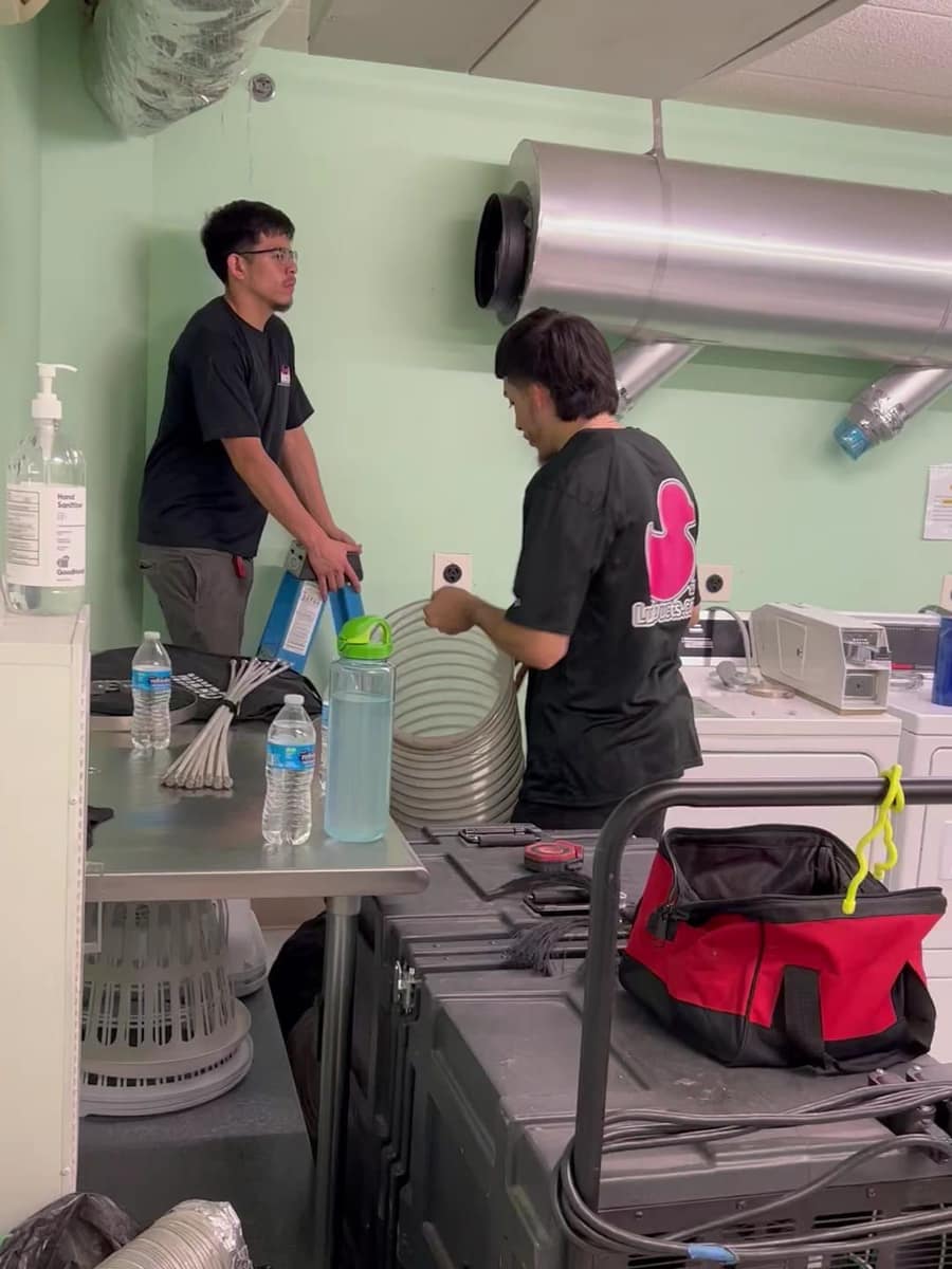 Two Luxe Ducts technicians in company shirts setting up in a utility room, with black negative air machine cases and coils of flexible duct in front of them