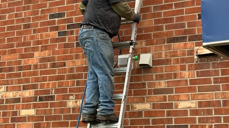 A technician on an extension ladder cleaning an exterior dryer vent on the brick wall of an apartment building